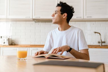Genç İspanyol adam evde mutfakta kahvaltı ederken kitap okuyor, masada oturuyor.