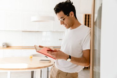 Gülümseyen genç İspanyol adam elinde kitapla evde duruyor.