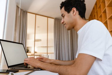 Düşünceli genç İspanyol adam evde masa başında otururken dizüstü bilgisayarda çalışıyor.