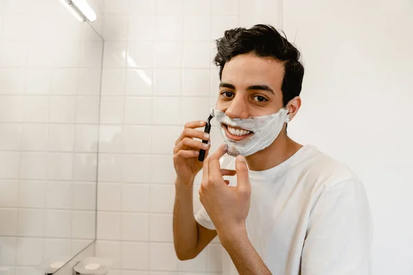 鏡の前で顔を剃っている若いヒスパニック系の男の笑顔室内 — ストック写真