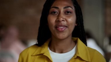 Sarı tişört giyen Afro-Amerikalı bir iş kadını ofiste dikilirken kameraya doğru bakıyor.