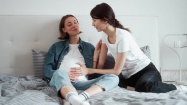 Bebek bekleyen mutlu lezbiyen kızlar yatak odasında otururken birbirleri ile konuşurken karnına dokunuyorlar.