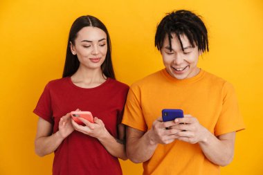 Sarı duvarda izole edilmiş, gülümseyen ve cep telefonu kullanan çok ırklı genç bir çift.
