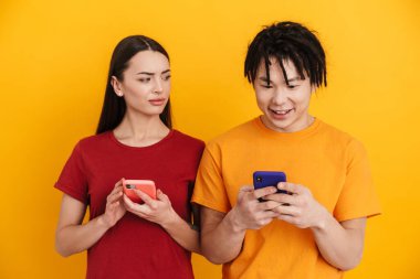Sarı duvarda izole edilmiş, poz veren ve cep telefonu kullanan çok ırklı genç bir çift.