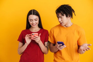 Sarı duvarda izole edilmiş, poz veren ve cep telefonu kullanan çok ırklı genç bir çift.