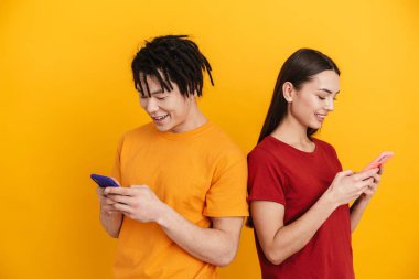 Sarı duvarda izole edilmiş, gülümseyen ve cep telefonu kullanan çok ırklı genç bir çift.