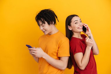 Sarı duvarda izole edilmiş cep telefonlarıyla konuşurken gülümseyen çok ırklı genç bir çift.