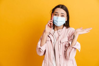 Sarı duvarda izole bir şekilde cep telefonuyla konuşurken yüzünde maske olan genç bir kadın.