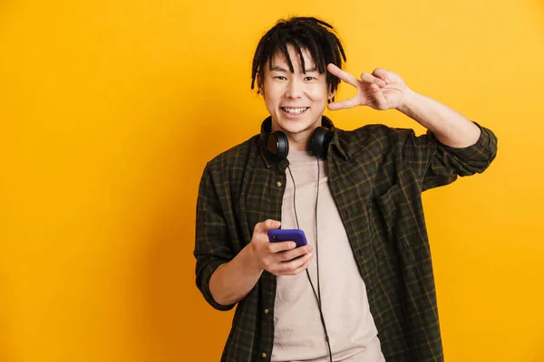 Uśmiechnięty Młody Azjata Trzyma Telefon Komórkowy Stojąc Nad Żółtym Tłem — Zdjęcie stockowe