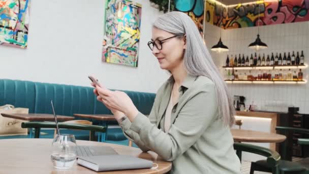 眼鏡をかけたビジネスマンの女性が 店内のカフェでノートパソコンを操作しながらスマホにタイプしている — ストック動画