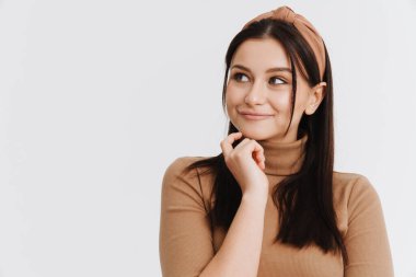 Düşünceli genç esmer kadın, günlük giysiler içinde beyaz arka planda dikiliyor, yan bakıyor.