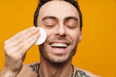 Üstsüz gülümseyen orta yaşlı esmer bir adamın yüzünü sarı arka planda pamukla temizlerken.