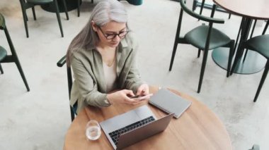 Gözlüklü bir iş kadınının en iyi görüntüsü, içerideki kafede laptopuyla çalışırken akıllı telefonuna mesaj yazması.