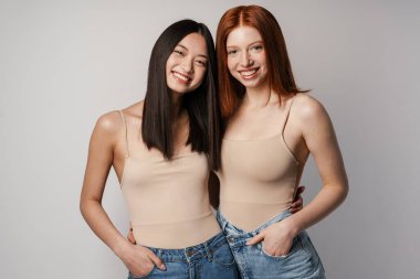 Çok ırklı iki kadın, beyaz duvardan izole edilmiş kameraya sarılıp gülümsüyor.