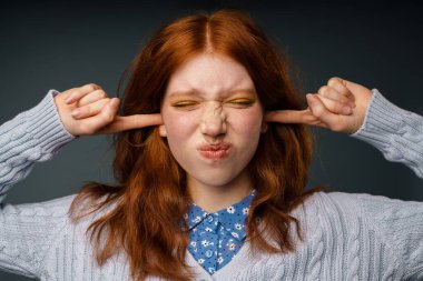 Gri arka planda kulaklarını tıkarken huysuz bir genç kadın.