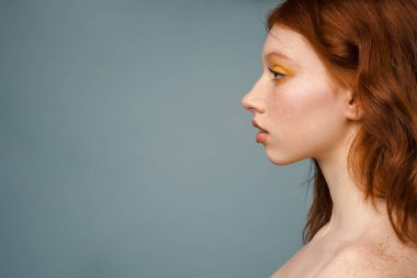 Tişörtsüz, kızıl beyaz kadın gri arka planda yalnız poz veriyor ve yan bakıyor.