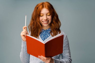 Gri arka planda izole edilmiş egzersiz kitabı ve kalemle poz veren kızıl saçlı genç kadın.