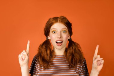 Turuncu arkaplanda parmaklarını yukarı kaldıran kızıl saçlı kadın şok oldu.