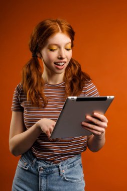 Turuncu arka planda izole edilmiş tablet bilgisayar kullanırken kuyrukları gülümseyen kızıl saçlı neşeli kadın.