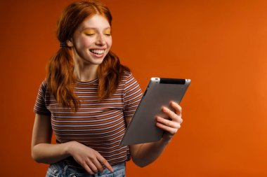 Turuncu arka planda izole edilmiş tablet bilgisayar kullanırken kuyrukları gülümseyen kızıl saçlı neşeli kadın.