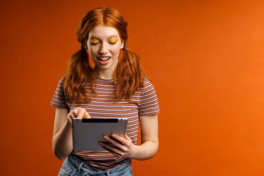 Turuncu arka planda izole edilmiş tablet bilgisayar kullanırken kuyrukları gülümseyen kızıl saçlı neşeli kadın.