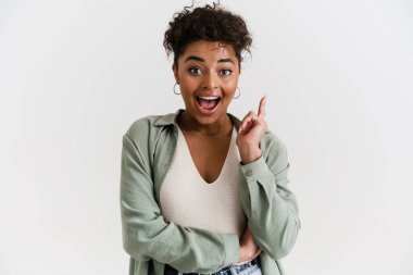 Smiling young african woman in casual wear standing over white wall, having an idea, pointing finger up