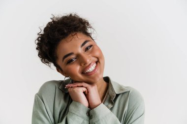 Smiling excited young african woman in casual wear standing over white wall, looking at camera, close up