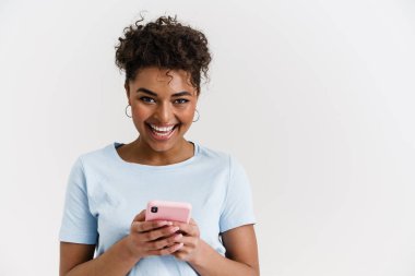Smiling happy young african woman in casual wear standing over white wall, holding mobile phone