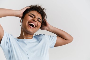 Happy casually dressed young african woman standing over white wall background, listening to music with earphones