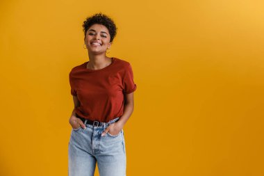 Happy casually dressed young african woman standing over yellow wall background, looking at camera