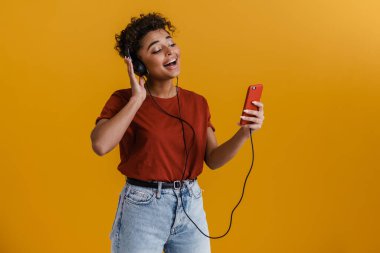 Happy casually dressed young african woman standing over yellow wall background, listening to music with earphones, holding mobile phone