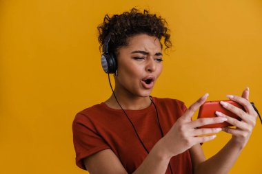 Surprised black woman listening music with headphones and cellphone isolated over yellow background