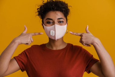 Black woman looking at camera while pointing finger at her face mask isolated over yellow background