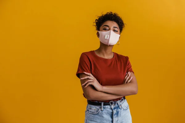 Young black woman in face mask posing with arms crossed isolated over yellow background