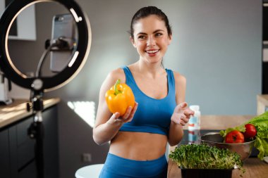 Ev mutfağında cep telefonundan selfie çekerken gülümseyen genç bir kadın.