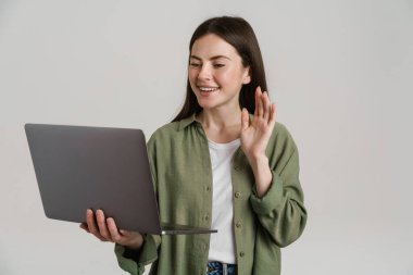 Mutlu, beyaz, sıradan bir kadın elinde dizüstü bilgisayarla arka planda el sallıyor.