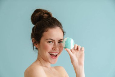 Mavi arka planda izole edilmiş banyo bombası pozu verirken gülümseyen yarı çıplak esmer kadın.