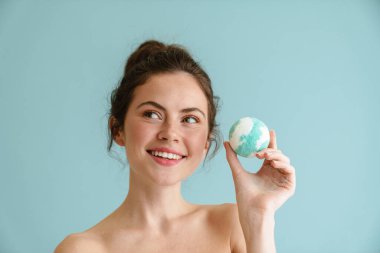 Mavi arka planda izole edilmiş banyo bombası pozu verirken gülümseyen yarı çıplak esmer kadın.