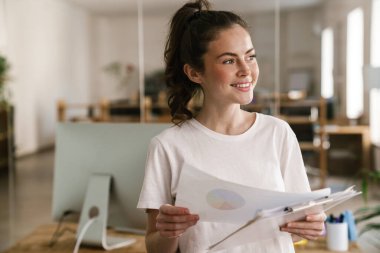 Gülümseyen, esmer, sıradan giyinmiş, büroda elinde belgelerle duran bir kadının fotoğrafını çek.