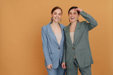 İki gülümseyen zarif kadın şık ceketler giyip birbirlerine yakın poz veriyorlar. Bej arka planda soyutlanmışlar.