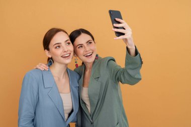 Sarı arka planda cep telefonundan selfie çekerken gülümseyen iki genç kadın.