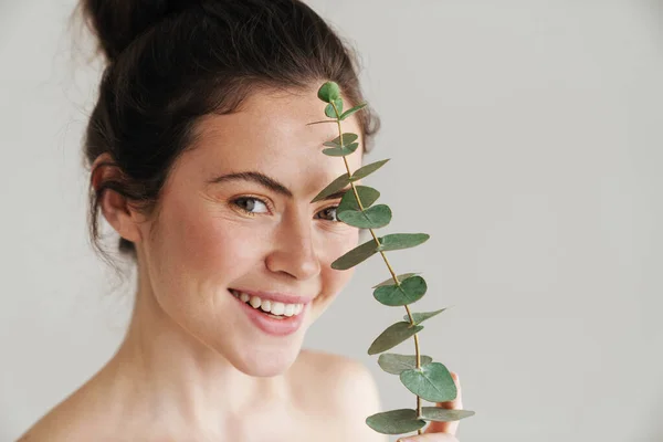 Half-naked brunette woman smiling while posing with eucalyptus isolated over while background