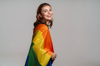 Gri arka planda izole edilmiş gökkuşağı bayrağıyla poz verirken gülümseyen mutlu güzel kadın.