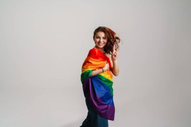 Gökkuşağı bayrağı takan neşeli genç kadın gri arka planda izole edilmiş barış işareti gösteriyor.