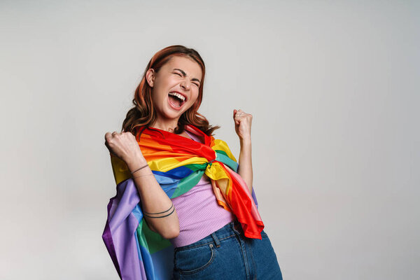 Возбужденная женщина делает жест победителя, позируя с радужным флагом, изолированным на сером фоне