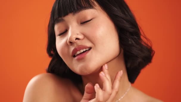 Detailní pohled na atraktivní asijské polonahá žena pózuje na kameru, zatímco se dotýká její tvář izolované přes oranžovou stěnu ve studiu