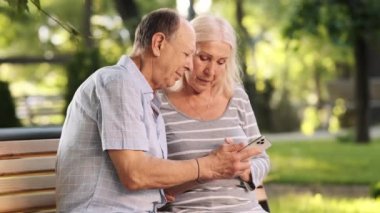 Memnun yaşlı bir çift parktaki bankta otururken cep telefonu kullanıyor.