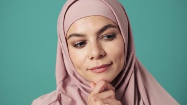 Pembe tesettürlü rüya gören bir Arap kadın mavi arka planda izole bir şekilde stüdyoda duran kameraya bakıyor.