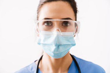 Tıbbi maskeli, koruyucu gözlüklü ve üniformalı güzel bayan doktor beyaz arka planda izole edilmiş, yakın plan.