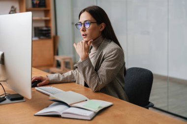 Ofiste otururken bilgisayar ve planlamacıyla çalışan gözlüklü çekici bir kadın.